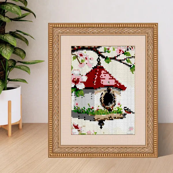 A framed color by number artwork depicting a birdhouse surrounded by blooming cherry blossoms, created with a mosaic pattern, displayed on a wooden surface next to a green plant.