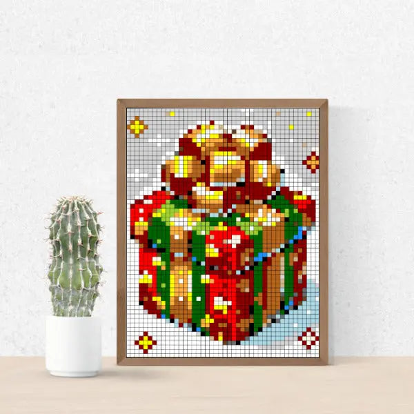 Framed Christmas pixel art color by number design showing a festive red and green gift box with a large golden bow, surrounded by snow and stars, displayed on a wooden surface next to a green cactus in a white pot.