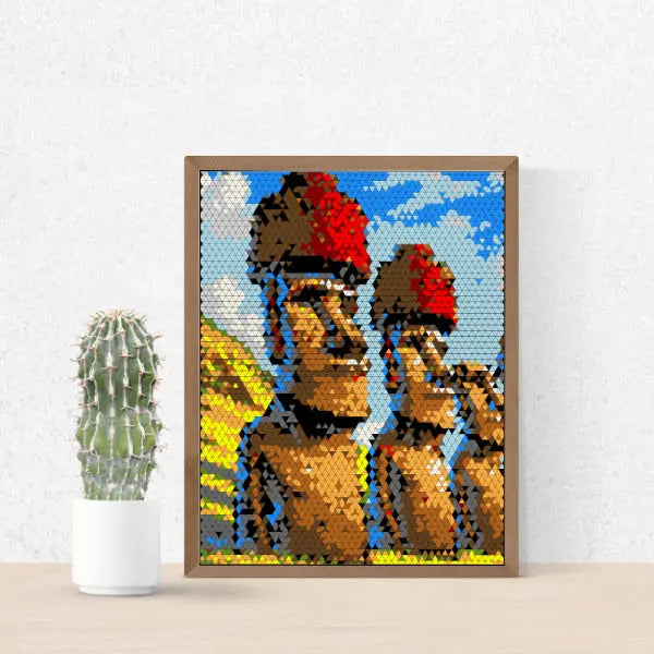A framed mosaic artwork on a wooden surface, depicting three Easter Island Moai statues with red topknots against a blue sky, created with small triangular shapes, next to a potted cactus.