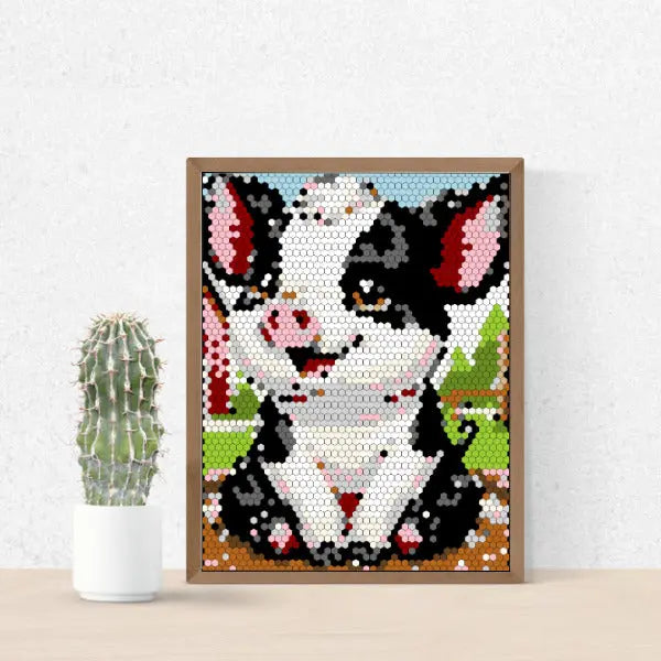 A framed color by number artwork depicting a cute piglet with black and pink spots, created with a hexagonal mosaic pattern. The artwork is displayed on a white surface next to a small potted cactus.