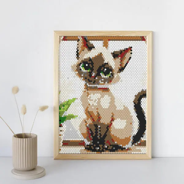 A framed color by number artwork depicting a siamese cat with blue eyes, created with a hexagonal mosaic pattern. The artwork is displayed on a light-colored surface next to a neutral-toned vase with a single dried decorative stem.