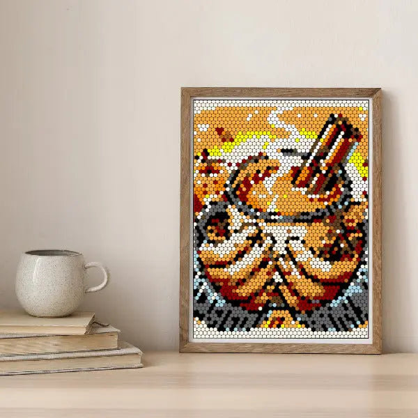 Framed Thanksgiving art color by number design showing a steaming hot mug of apple cider with a cinnamon stick, rendered in orange and brown tones, displayed on a wooden table next to a stack of books and a ceramic mug.