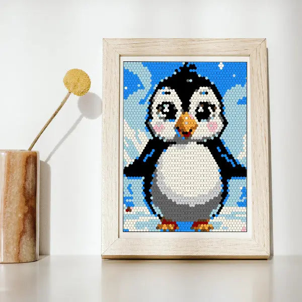 A framed color by number artwork depicting an adorable baby penguin with rosy cheeks against a snowy and blue sky background, created with a hexagonal mosaic pattern. The artwork is displayed on a white surface next to a neutral-toned vase with a single dried decorative stem.