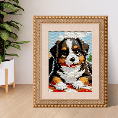 A framed color by number artwork depicting a Bernese Mountain Dog, created with a hexagonal mosaic pattern, resting on a red blanket. The artwork is displayed on a wooden surface next to a green plant.